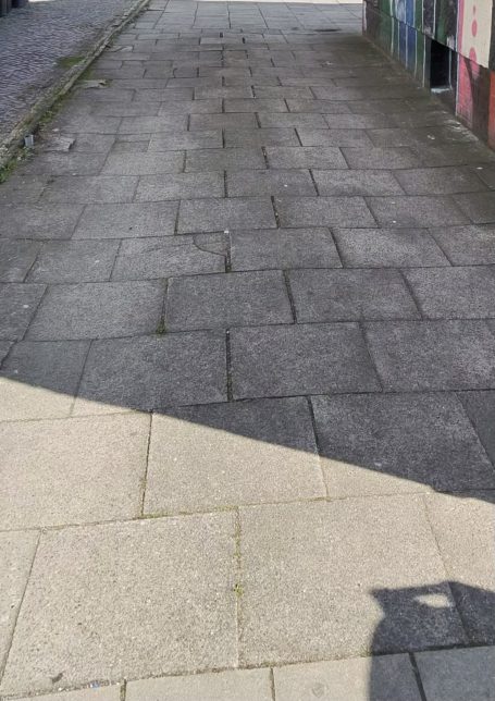 Gepflasterter Gehweg mit Schatten und bunten Wänden im Hintergrund.